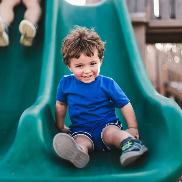 Child Enjoying a Slide A young child with curly hair wearing a blue shirt and shorts is sitting on a green slide, smiling happily. Another child is slightly behind, going down the slide in the background.