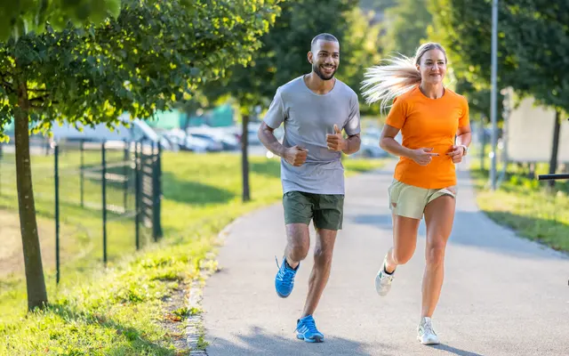 A man and a woman jogging together on a paved path lined with trees, wearing athletic clothing. The man is in a gray shirt and shorts, while the woman is dressed in an orange top and light shorts. They both appear to be enjoying their run.
