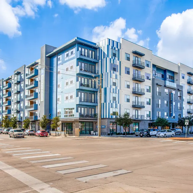 A modern multi-story apartment building with balconies and a mix of blue and gray facade, located at a busy intersection with vehicles on the street.
