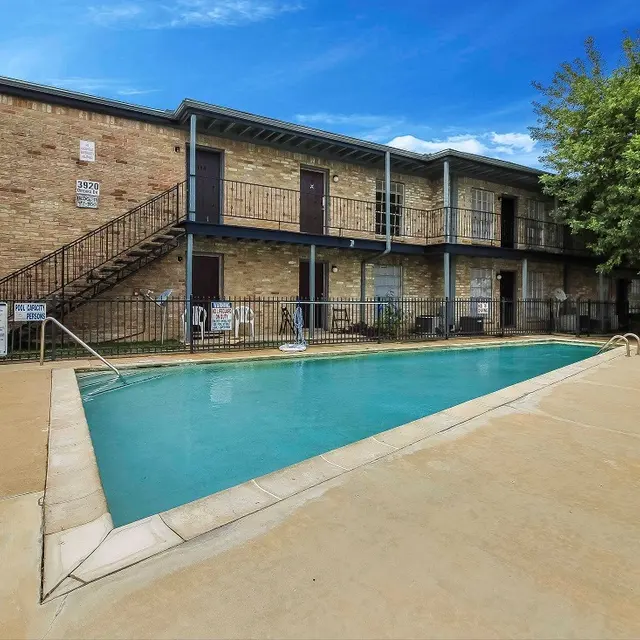 An exterior view of a two-story apartment complex with a swimming pool in the foreground. The building features brick walls and a staircase on the left side, with several doors visible on the balcony. Lush greenery surrounds the area, and the sky is bright with scattered clouds.
