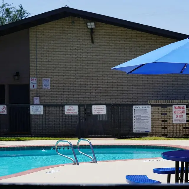 View of a swimming pool area with a blue umbrella, chairs, and a building in the background.