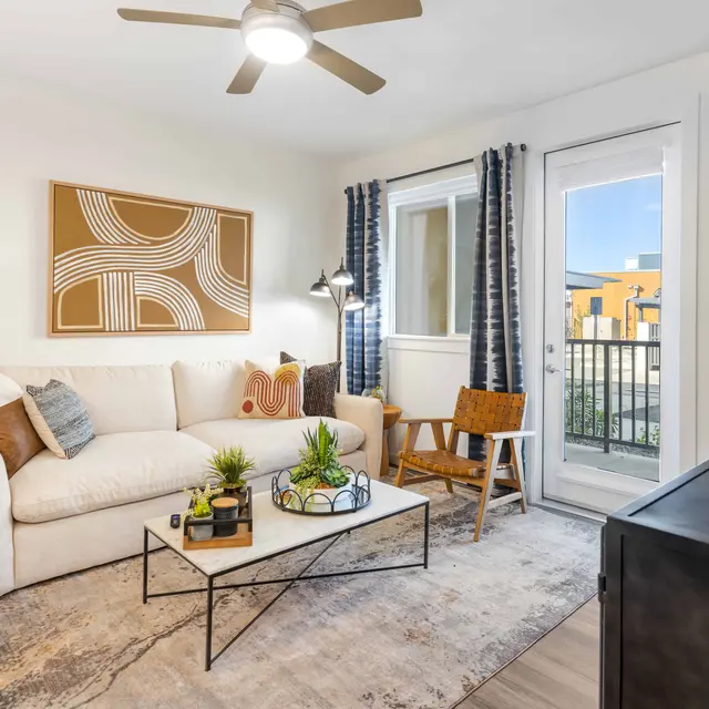 A modern living room featuring a white sofa with decorative pillows, a glass coffee table with plants, a wooden side table, and large windows with curtains. A contemporary art piece is on the wall.