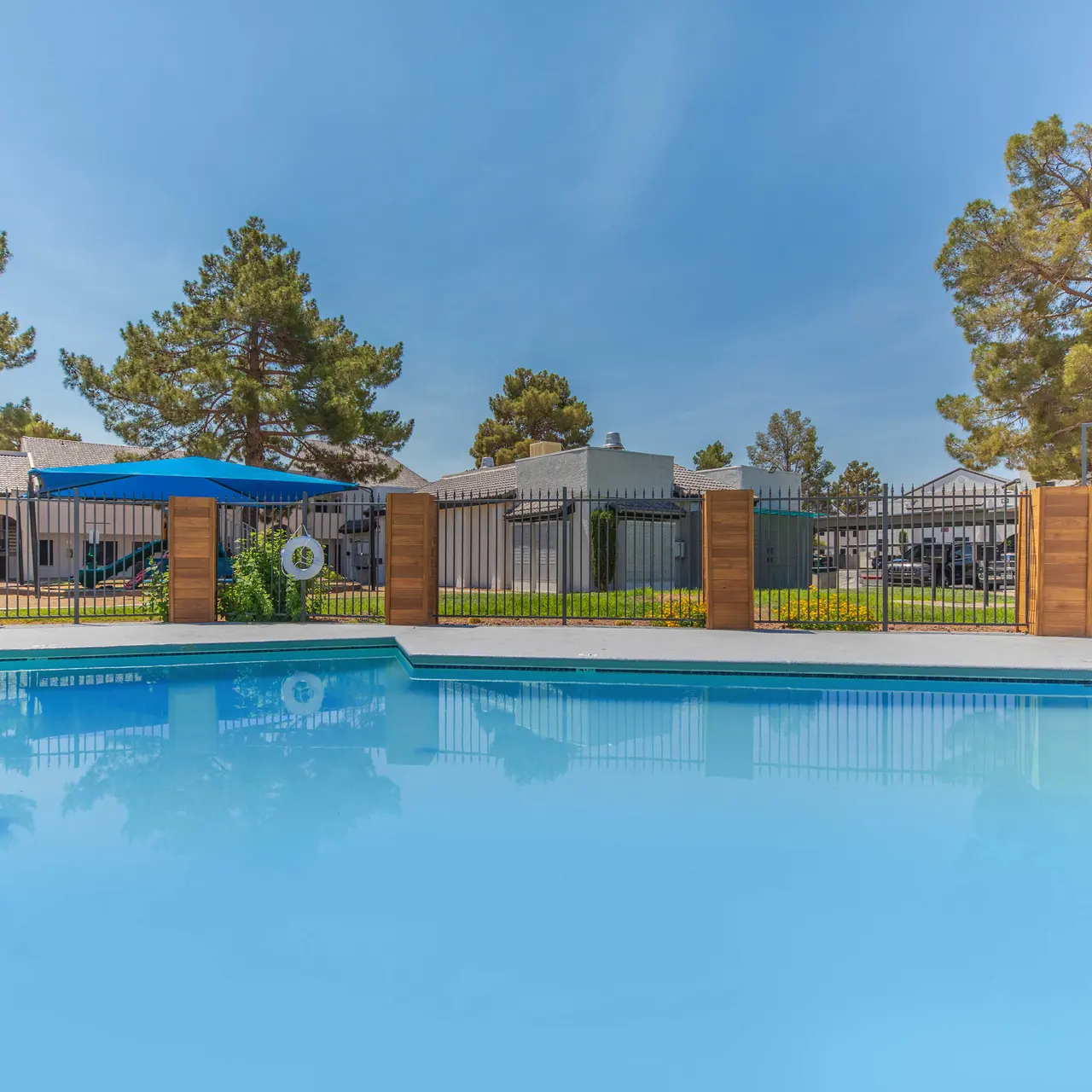 A swimming pool with clear blue water surrounded by a landscaped area with trees and a fenced perimeter. Sun loungers and umbrellas are visible in the background, creating a relaxing atmosphere.