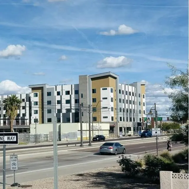 A modern apartment building with a combination of white and yellowish exterior, located beside a busy road with vehicles. The sky is clear with some clouds, and there are palm trees in the foreground.