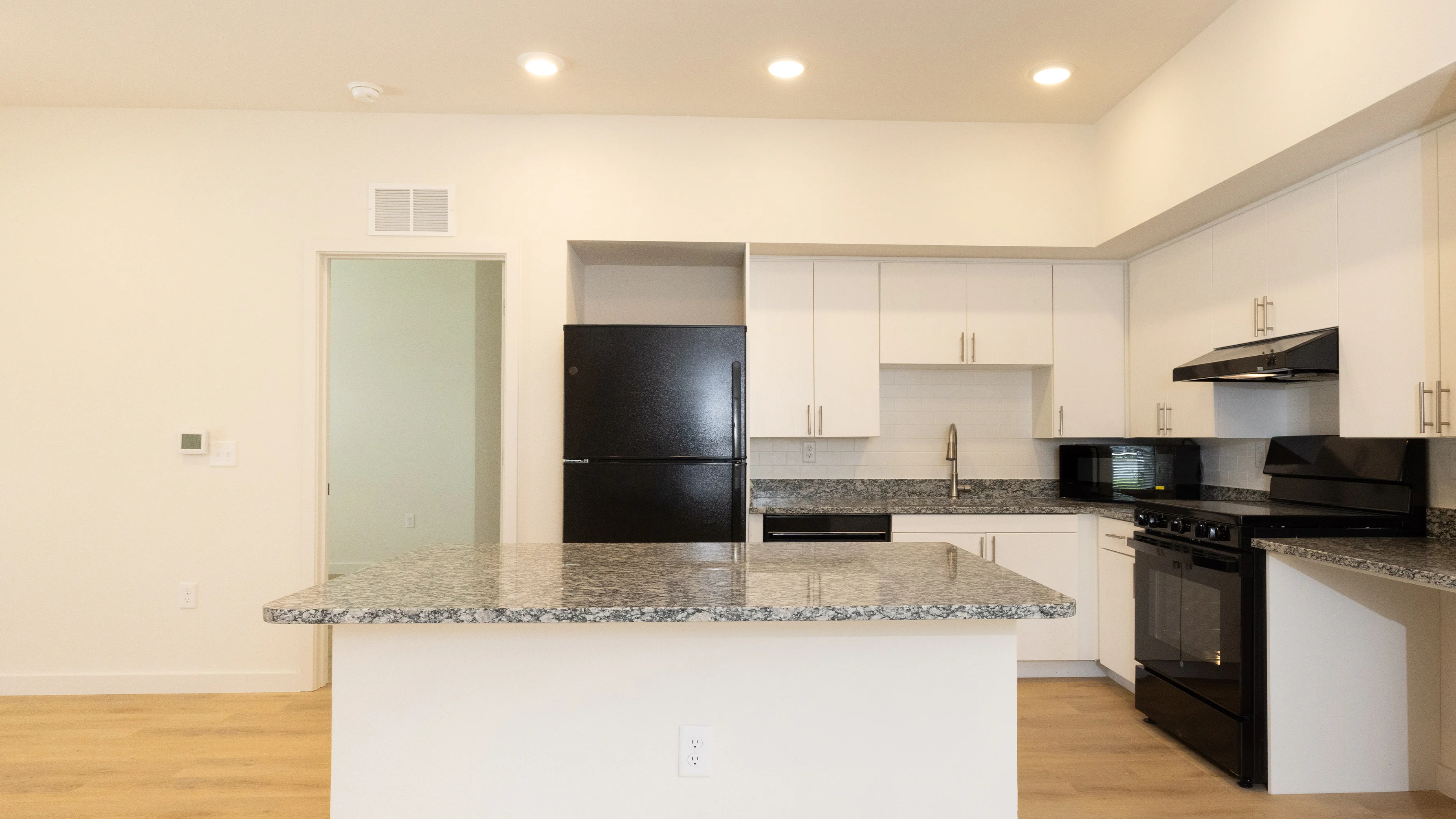 A modern kitchen interior showcasing a spacious layout with white cabinetry, granite countertops, and stainless steel appliances including an oven, microwave, and refrigerator. The flooring is light wood, and the walls are painted in a neutral tone, enhancing the bright and airy atmosphere.