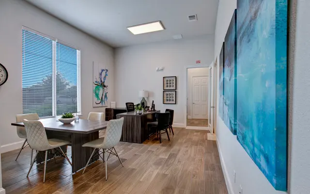 A contemporary office interior featuring a large table, chairs, a desk, and colorful artwork on the walls. Large windows allow natural light to fill the space.