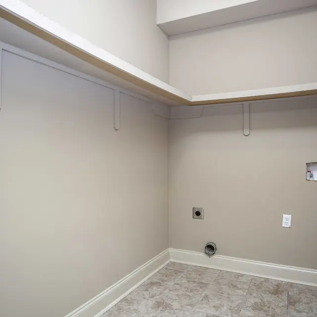 An empty laundry room with light beige walls and tiled flooring. There are shelves along the walls and plumbing connections visible.