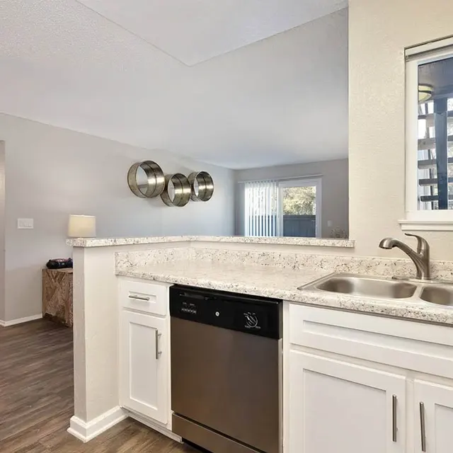 A modern kitchen featuring white cabinets, stainless steel appliances, and a granite countertop with a double sink. A dining area with a glass table and a few chairs is visible. The background shows a living space with large windows allowing natural light.