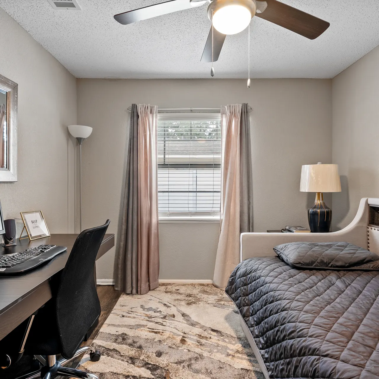 A modern, cozy room featuring a workspace with a desk and office chair, a single bed with a dark comforter, a large window with curtains, and a decorative wall art piece.