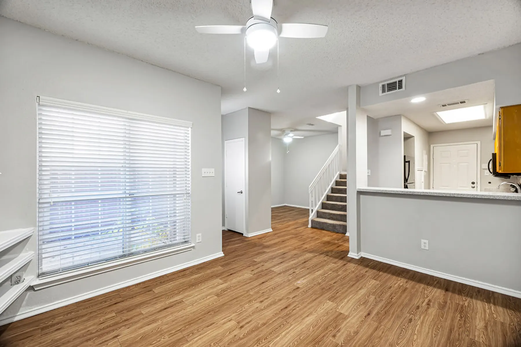Modern Living Room Interior Spacious living room area with light-colored walls and wooden flooring, featuring a ceiling fan, windows with blinds, and an open layout towards the kitchen and staircase.