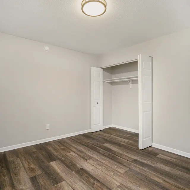 A spacious, empty bedroom with light-colored walls and wooden flooring. There is an open closet with sliding doors on the left side of the room. A doorway is visible on the right side, leading to another space.
