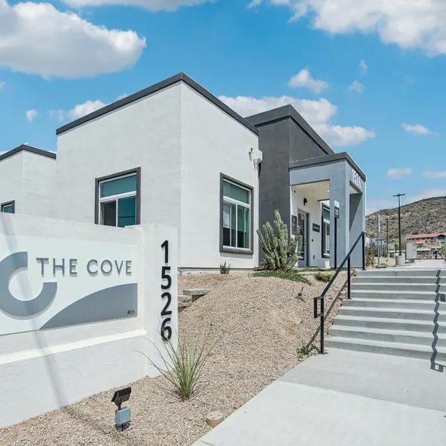 A modern building complex with a sign that reads 'The Cove' and the number '1526'. The structure has a contemporary design with large windows and a path leading up to the entrance. Surrounding the building is a landscaped area with desert vegetation and a clear blue sky.