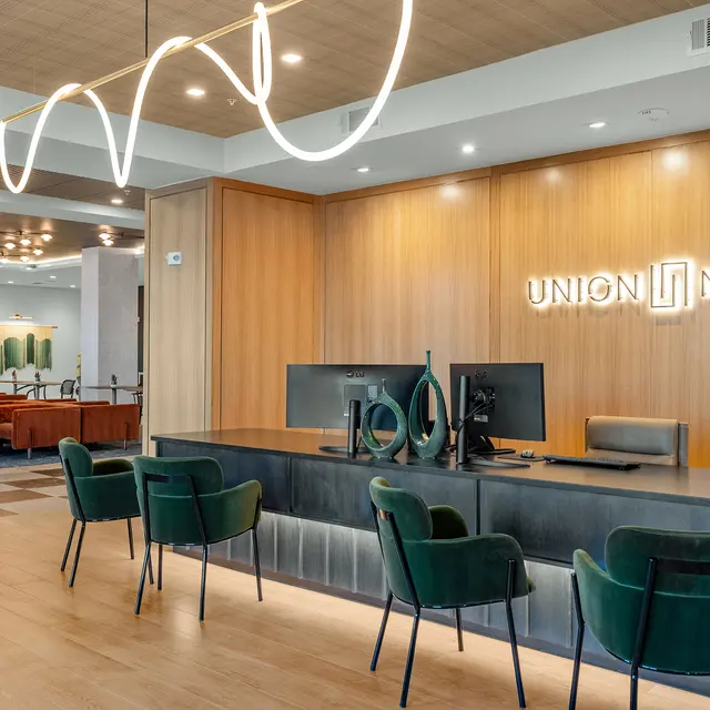Modern reception area with wooden paneling, a sleek desk, computer monitors, and green upholstered chairs.