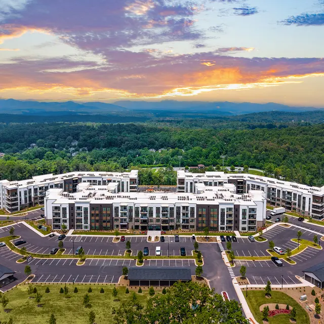 Aerial view of a modern apartment complex with a spacious parking area and surrounding greenery under a colorful sunset sky.