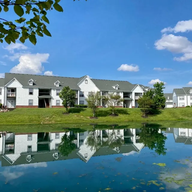 A serene apartment complex reflected in a pond, surrounded by green grass and trees under a blue sky with fluffy white clouds.