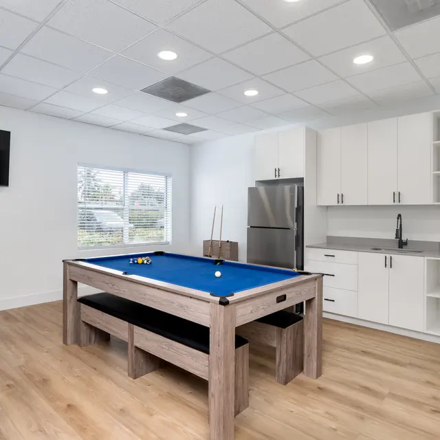 A bright, modern game room featuring a blue pool table, a comfortable grey bean bag, and a stainless steel kitchen area with cabinets and a refrigerator.