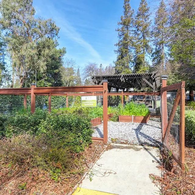 A well-maintained park entrance with a wooden fence and a path leading into a green area surrounded by trees and bushes.