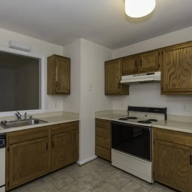 A kitchen with wooden cabinets, a white stove, and a refrigerator, with a countertop and sink area. The walls are painted in a light color, and there is a window above the sink.