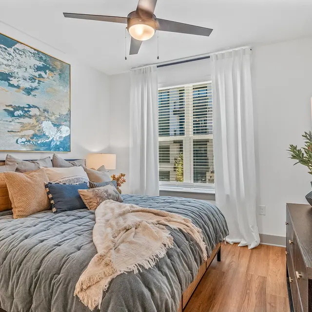A stylish modern bedroom featuring a large painting above a bed adorned with various pillows and a decorative throw, with a fan overhead and large window with sheer curtains.