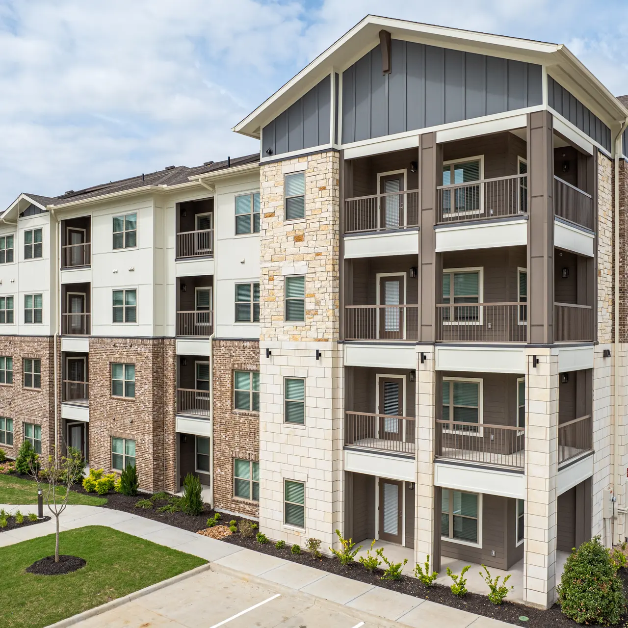 A modern apartment building featuring a mix of stone and siding exteriors, with balconies on the upper floors and a landscaped area in front.
