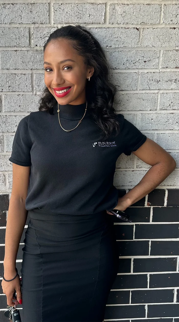 A smiling woman posing against a brick wall, wearing a black shirt and a black skirt.