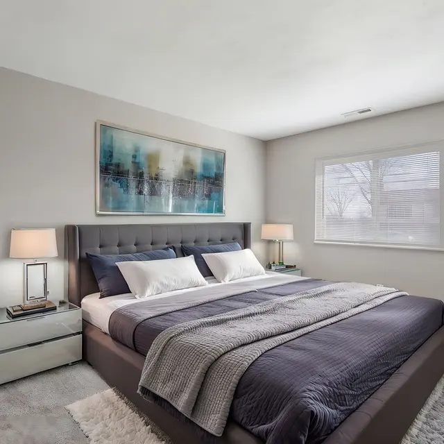 A modern bedroom featuring a gray upholstered bed with white and navy bedding, two bedside lamps, and a large window letting in natural light. There is a potted plant in the corner and abstract artwork on the wall.