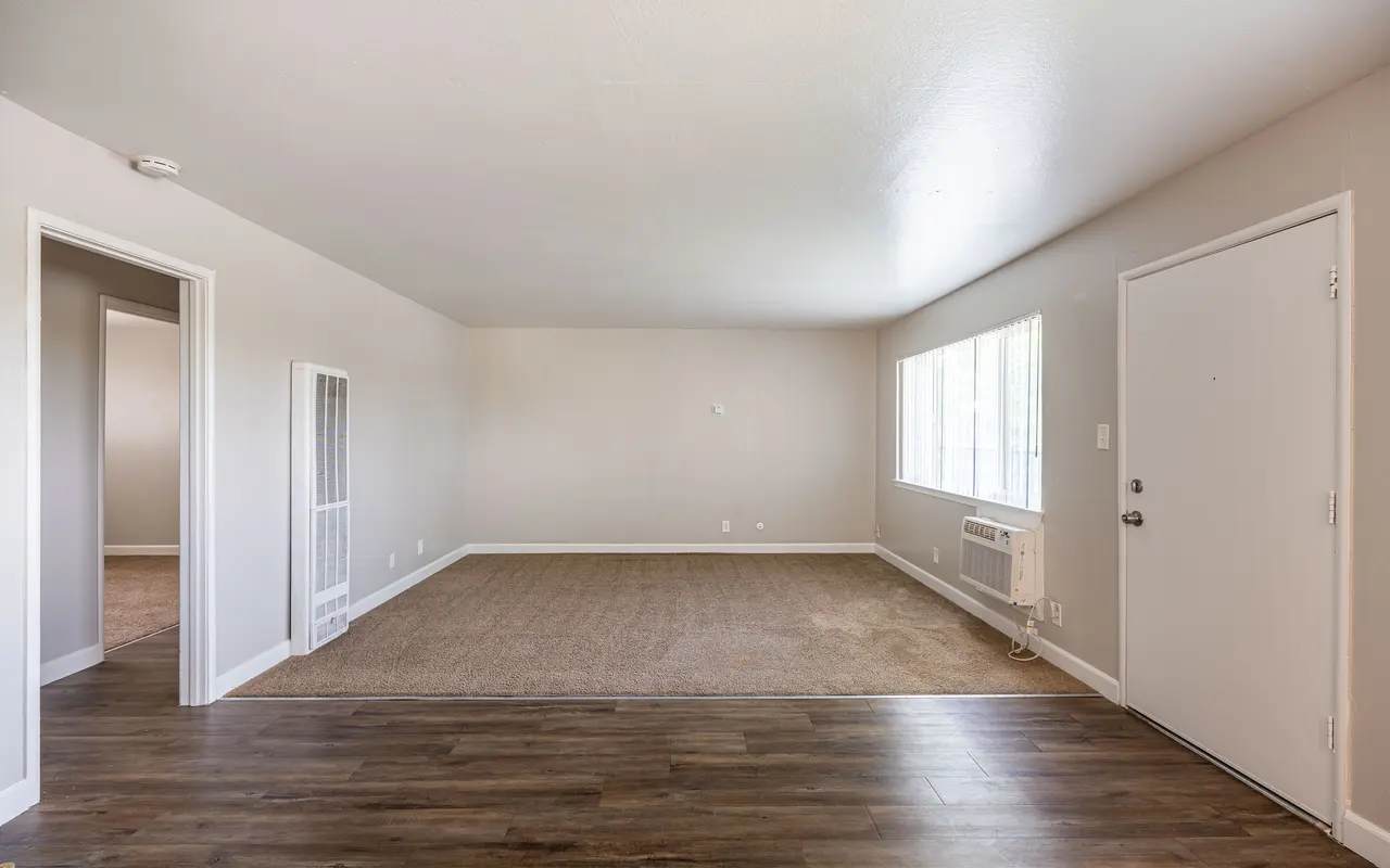 A spacious, empty living room with light-colored walls and large windows. The flooring features a combination of wood and carpet. There is an entrance door on the right and a small air conditioning unit beneath the window.