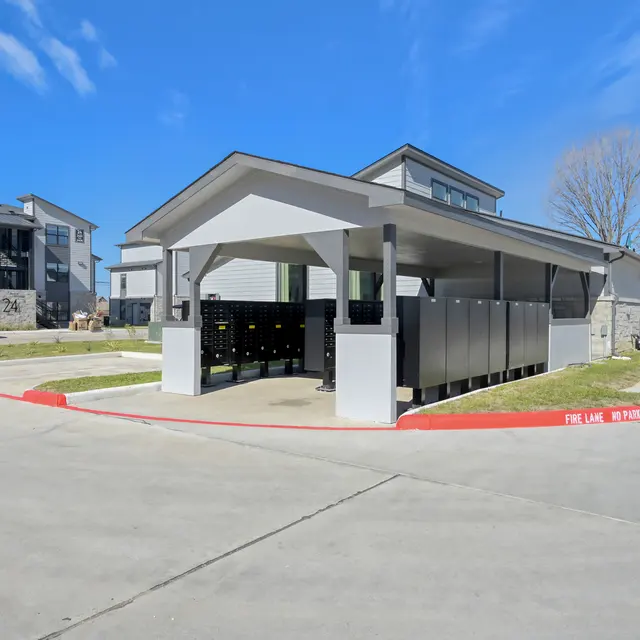 A covered parking structure with storage lockers, surrounded by an asphalt lot and modern apartment buildings in the background under a blue sky.