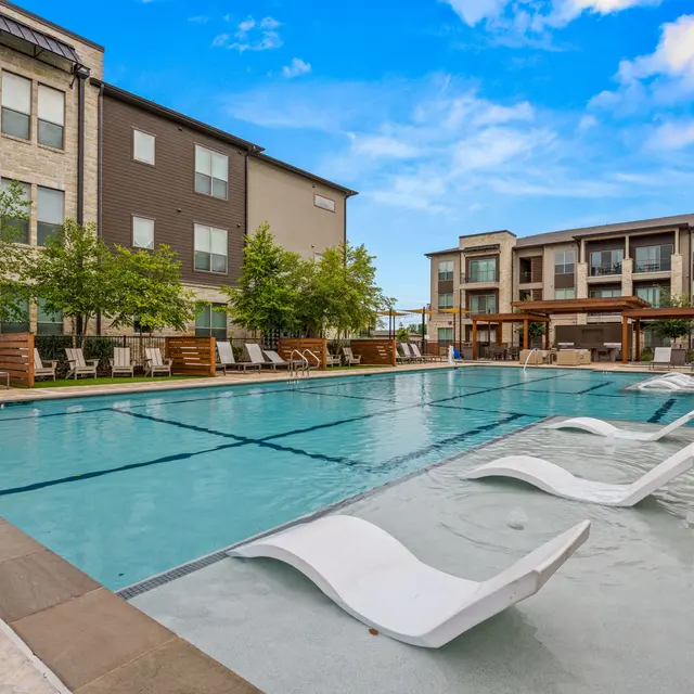 A spacious pool area surrounded by modern apartment buildings. Lounge chairs are positioned around the pool with trees and greenery nearby, under a bright blue sky.