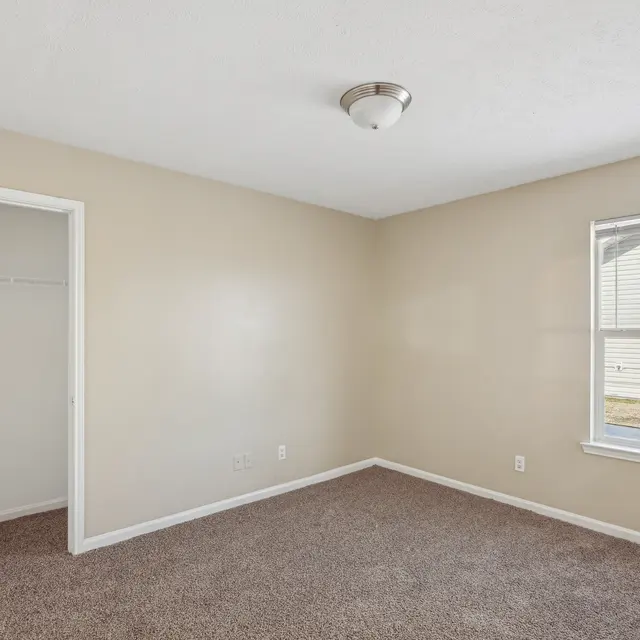 A vacant bedroom with beige walls and carpet flooring. It features a window letting in natural light, and a closet with open doors. Lighting fixture on the ceiling.