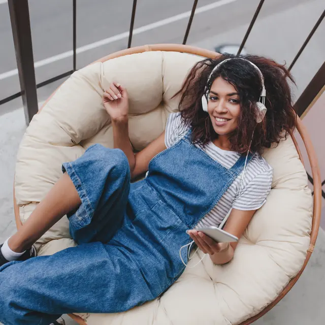 A young woman with curly hair sitting in a large, round chair on a balcony. She is wearing denim overalls and a striped shirt, listening to music through headphones, while holding a smartphone. The balcony has a view of a street and other buildings.