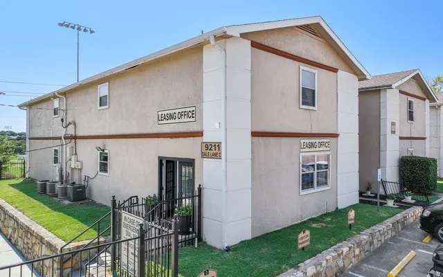 Leasing Office Exterior Exterior view of a leasing office building featuring light-colored walls, signage indicating 'Leasing Office', and a fenced area with greenery and parking spaces.