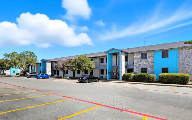 Exterior view of a two-story apartment complex featuring light blue and gray stone exteriors, with trees and cars parked in front. The sky is clear with some fluffy clouds.