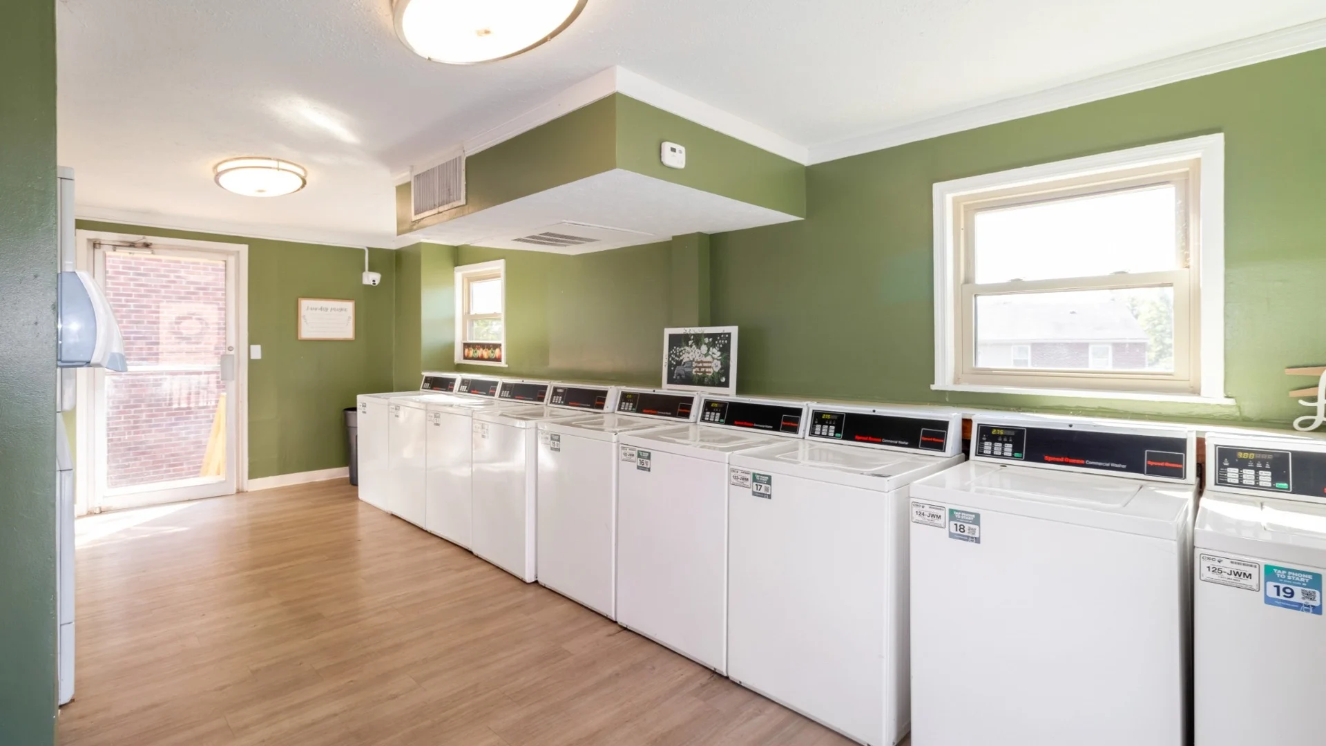 Bright laundry room with green walls and several white washing machines, featuring wooden flooring and natural light from a window.
