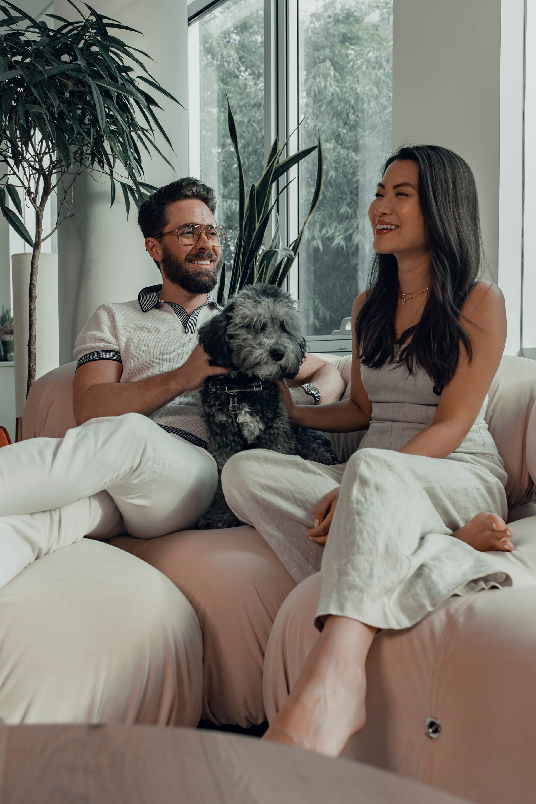 A man and a woman sitting on a couch, smiling and enjoying the company of a small dog. The room is bright with large windows and indoor plants.