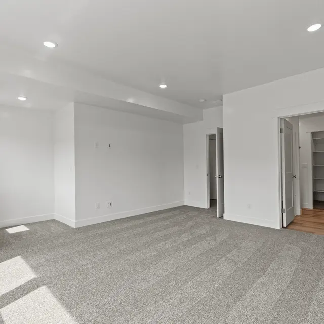 A spacious, empty room with light gray carpet flooring and white-painted walls. There are two doors leading to other spaces, and ample natural light coming in from windows.