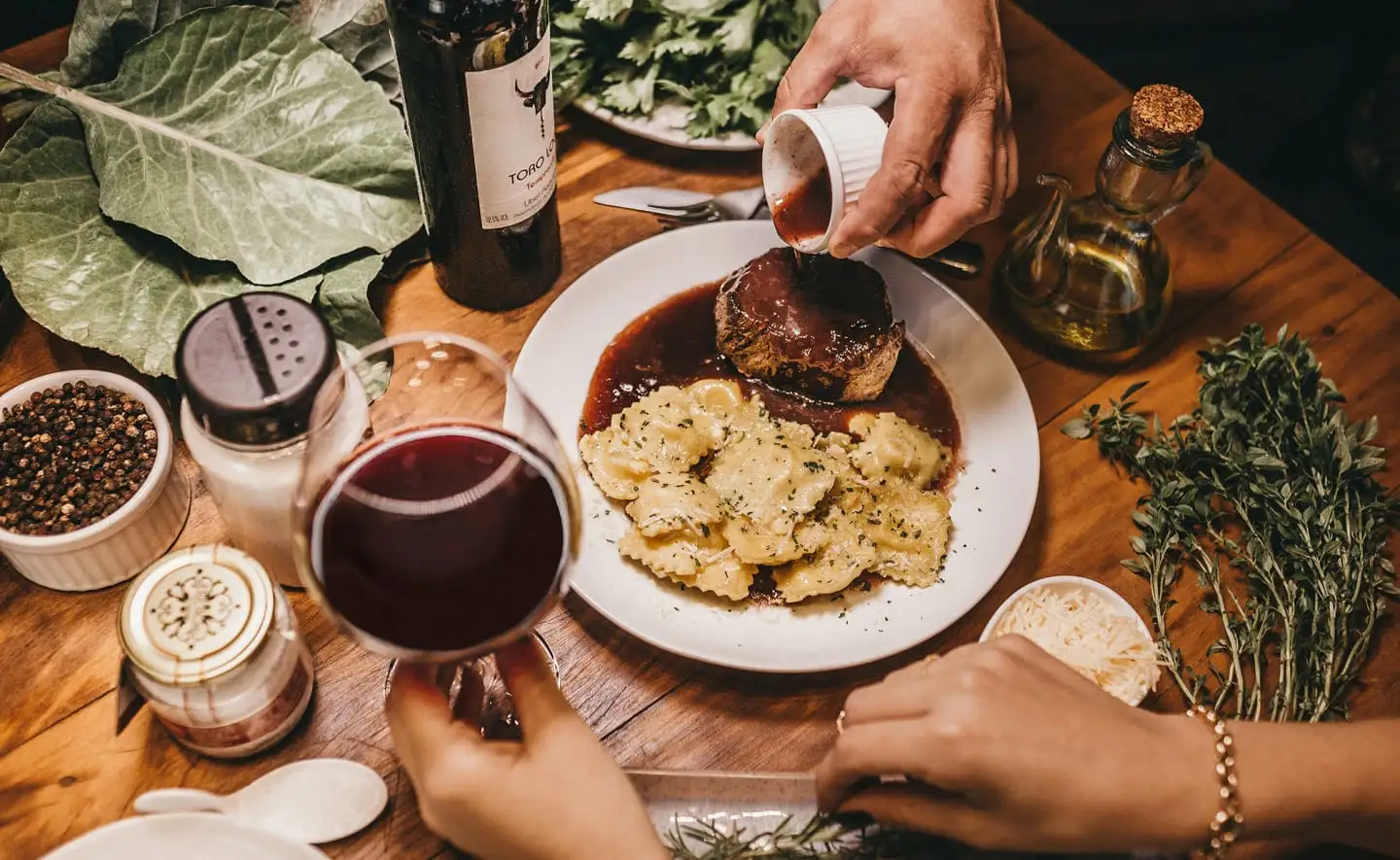 An overhead view of a dining table set with a plate of ravioli and meat, surrounded by glasses of red wine, herbs, and condiments, creating a cozy dining atmosphere.