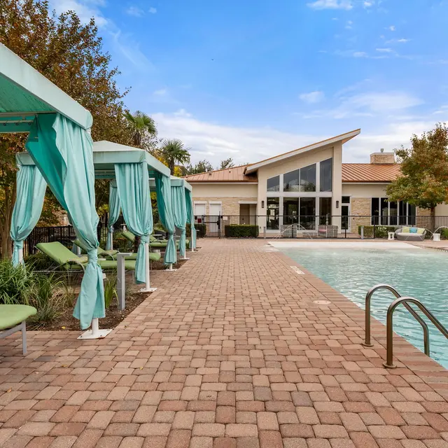 A view of a swimming pool area with cabanas and lounge chairs beside it, surrounded by landscaped greenery.