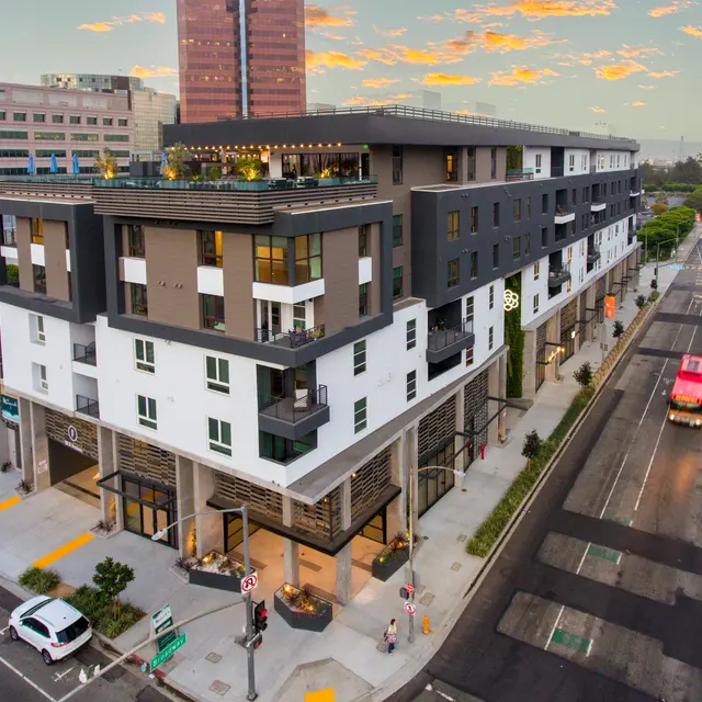 Urban Modern Apartment Building A modern multi-story apartment building with a flat roof, situated on a city street. The ground level features commercial spaces with glass facades, and there are cars parked on the street. Skyscrapers are visible in the background against a cloudy sky.