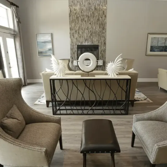 A stylish modern living space featuring two upholstered armchairs facing a small leather ottoman, with a decorative table behind them. The room is well-lit with large windows and has neutral-toned walls and flooring. Decorative accents and artwork are displayed on the walls.