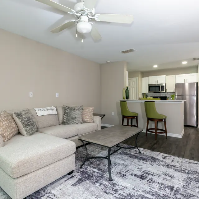 A stylish modern living room featuring a beige sectional sofa with decorative pillows, a coffee table, and a rug. A kitchen area with white cabinetry and stainless steel appliances can be seen in the background.