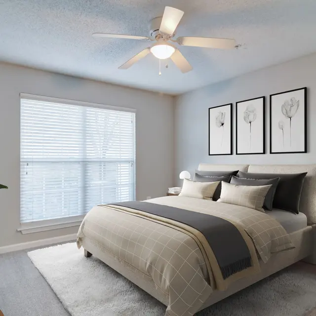 A modern bedroom featuring a queen-sized bed with gray bedding, decorative pillows, and a stylish headboard. The room has a ceiling fan and a soft rug under the bed. A bedside table with a lamp and a small plant is visible, along with three framed botanical prints on the wall. Natural light streams in through a window with blinds.