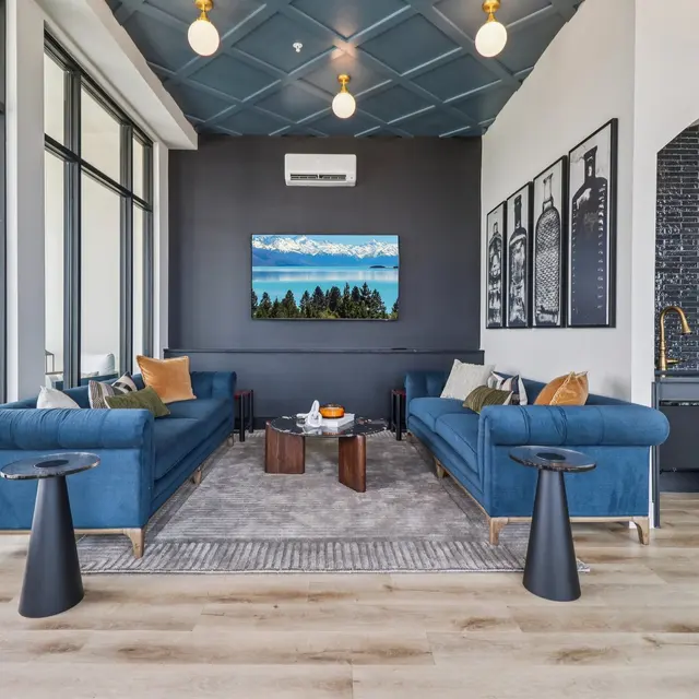 A modern living room with two blue sofas, a large rug, and a wall-mounted TV displaying a scenic view. The room features large windows, pendant lights, and a sleek kitchen area in the background.