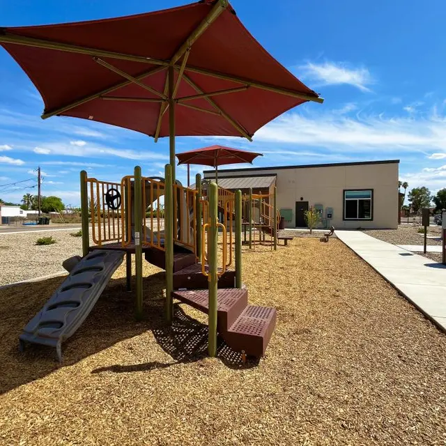 Sunny Playground with Shade Canopies A playground structure with two red shade canopies, accompanied by a slide and brown wood chips covering the ground. A building is visible in the background under a blue sky with scattered clouds.