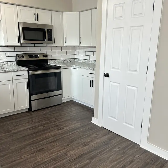 A modern kitchen featuring white cabinets, stainless steel appliances, and a tiled backsplash in a light color. The flooring is dark wood, and there is a door on the right side leading to another room or space.