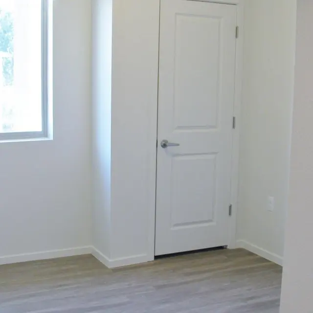An empty room featuring light-colored walls and a door. A window allows natural light to enter, illuminating the wooden flooring.