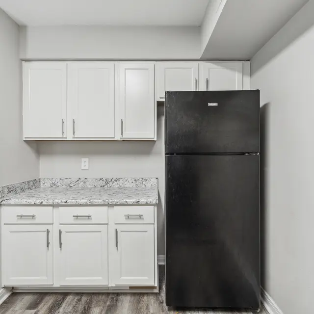Modern kitchen corner with black refrigerator, white cabinets, and a granite countertop. Light gray walls add brightness.