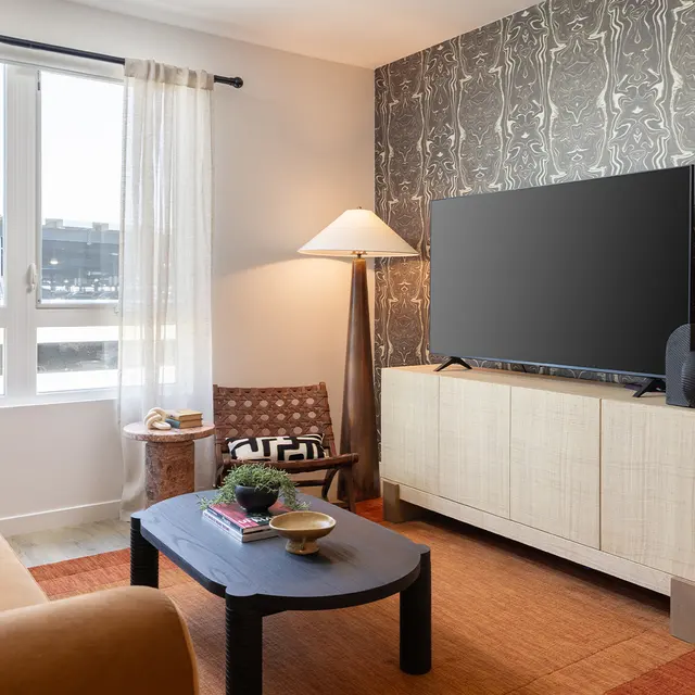 A modern living room featuring a stylish design with a large television, a light-colored sofa, a decorative coffee table, and greenery. The walls are adorned with a patterned wallpaper, and large windows allow natural light to fill the space.