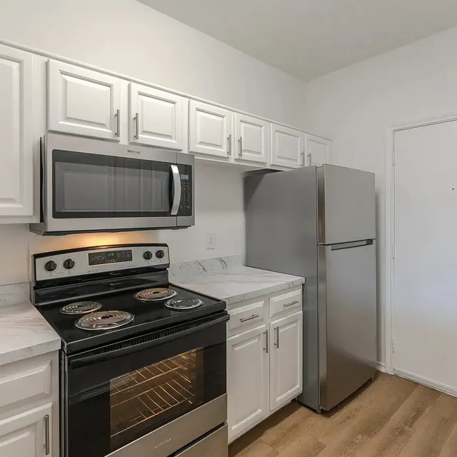 A modern kitchen featuring white cabinets, stainless steel appliances, and a marble countertop. The layout includes an electric stove, a built-in microwave, and a refrigerator with a door leading outside.