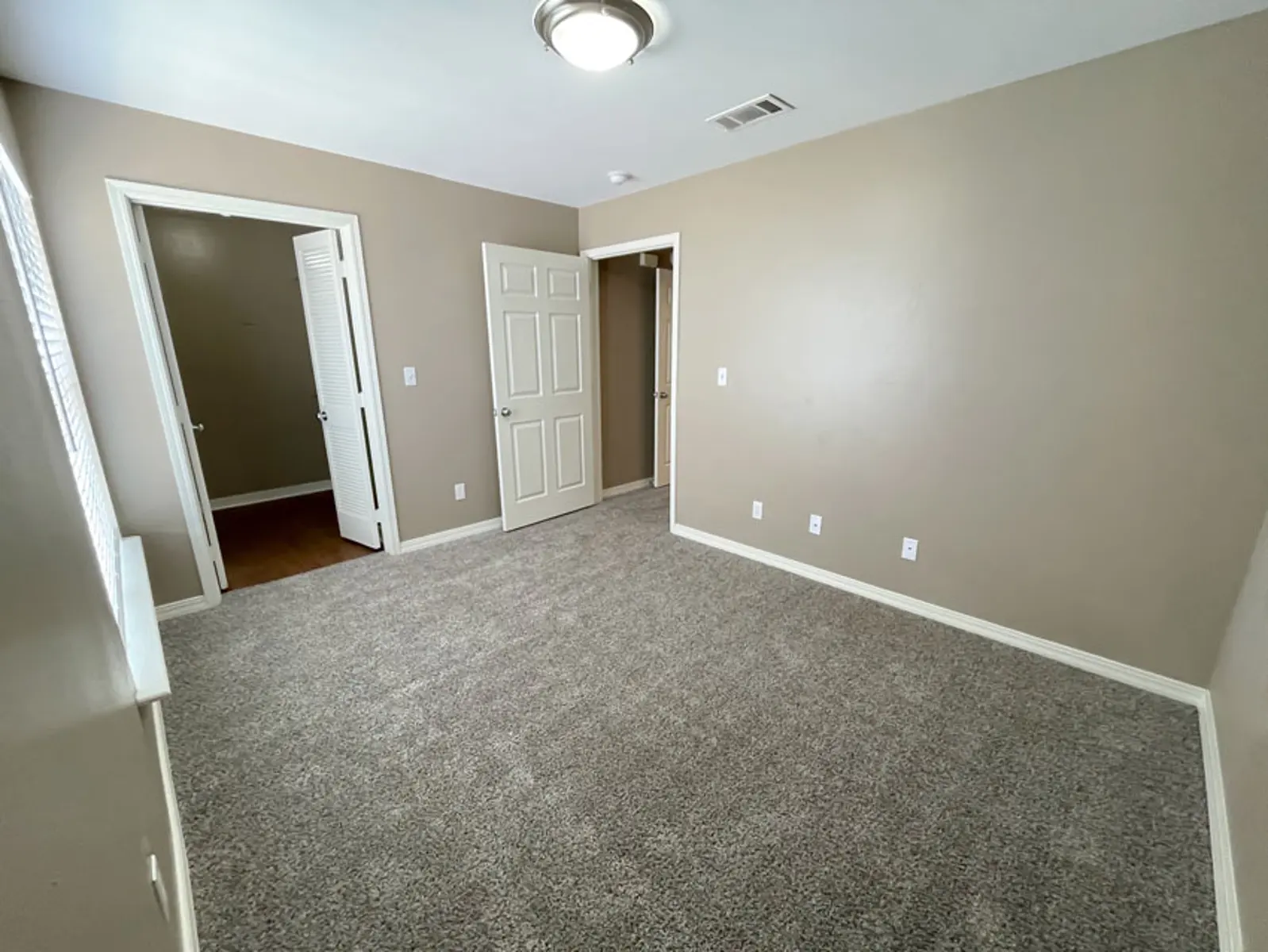 A vacant bedroom with light beige walls and carpeted floor, featuring two doors leading to other areas and a ceiling light fixture.